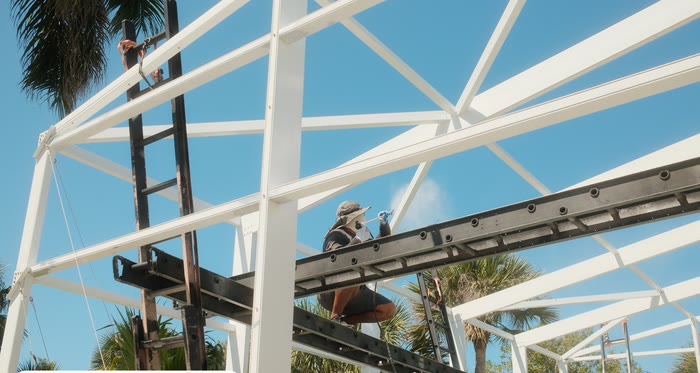A painter up on the frame spraying a bare cage white, palms behind.