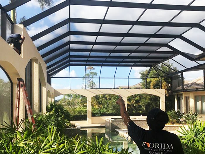 A painter in a branded shirt spraying the inside of a screened enclosure beside a pool.