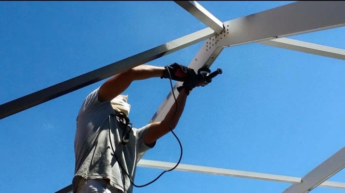 Close up of a spray gun coating a cage beam, palms and blue sky behind.