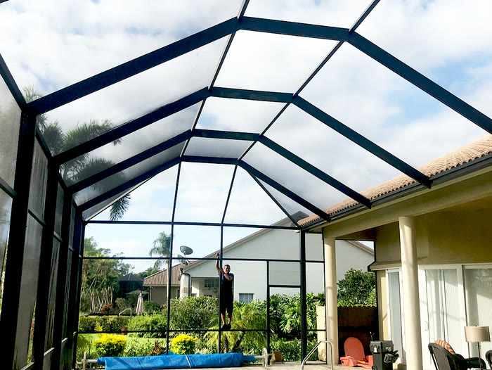 A white screened enclosure over a pool, with a yellow house behind it.