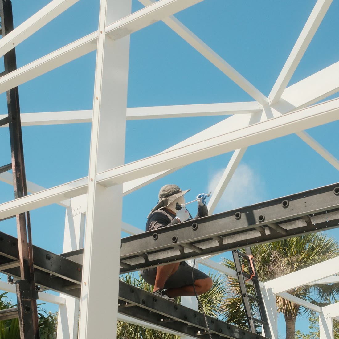 A painter up on the frame, spraying a pool cage beam white against a blue sky.